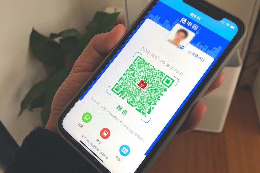 You view a hand holding a smartphone closeup with a green QR code in the centre of the screen against a blue background.